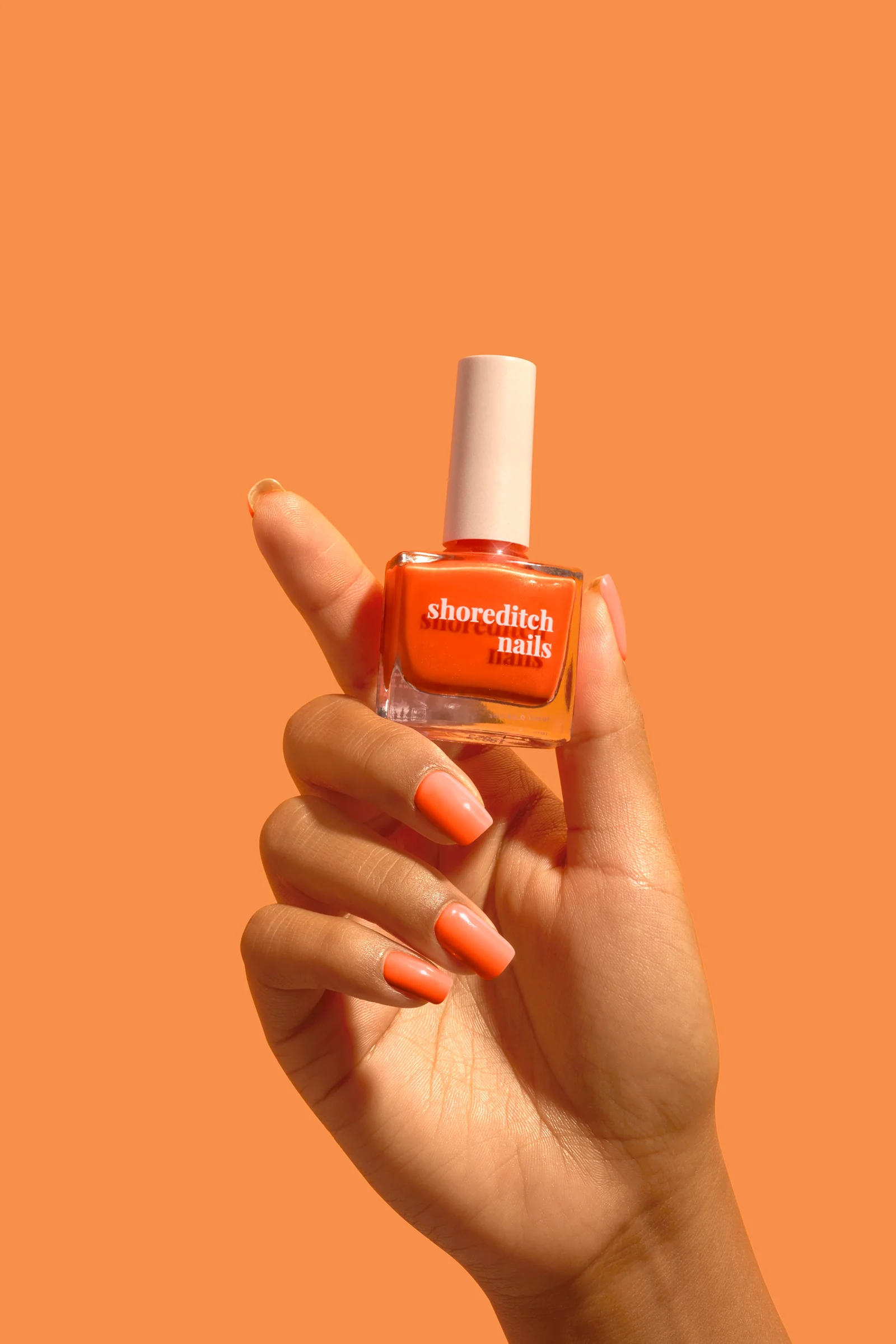 Hand with orange-painted nails holding an orange Shoreditch Nails polish bottle against a matching orange background.