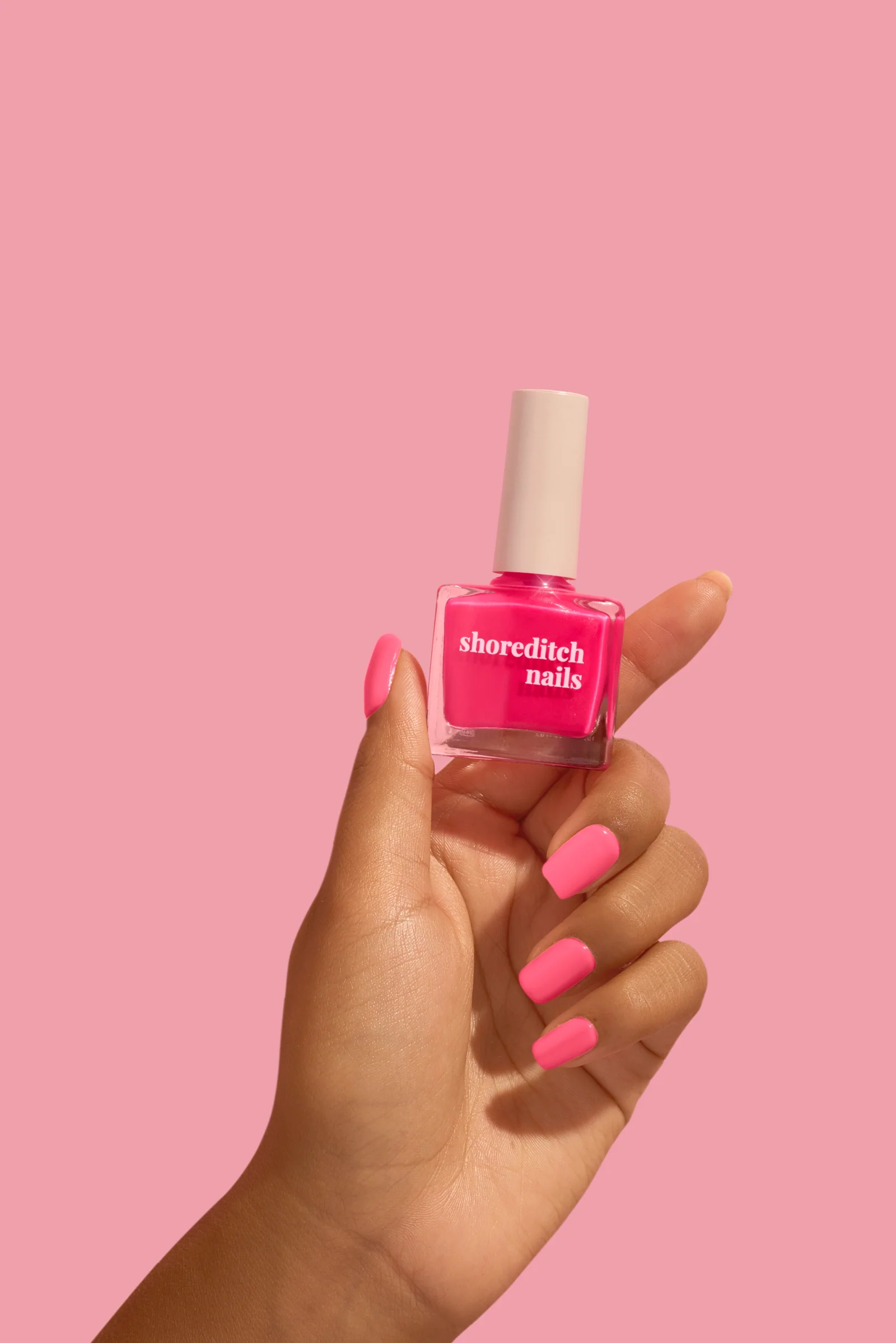 Hand with pink-painted nails holding a pink Shoreditch Nails polish bottle against a matching pink background.