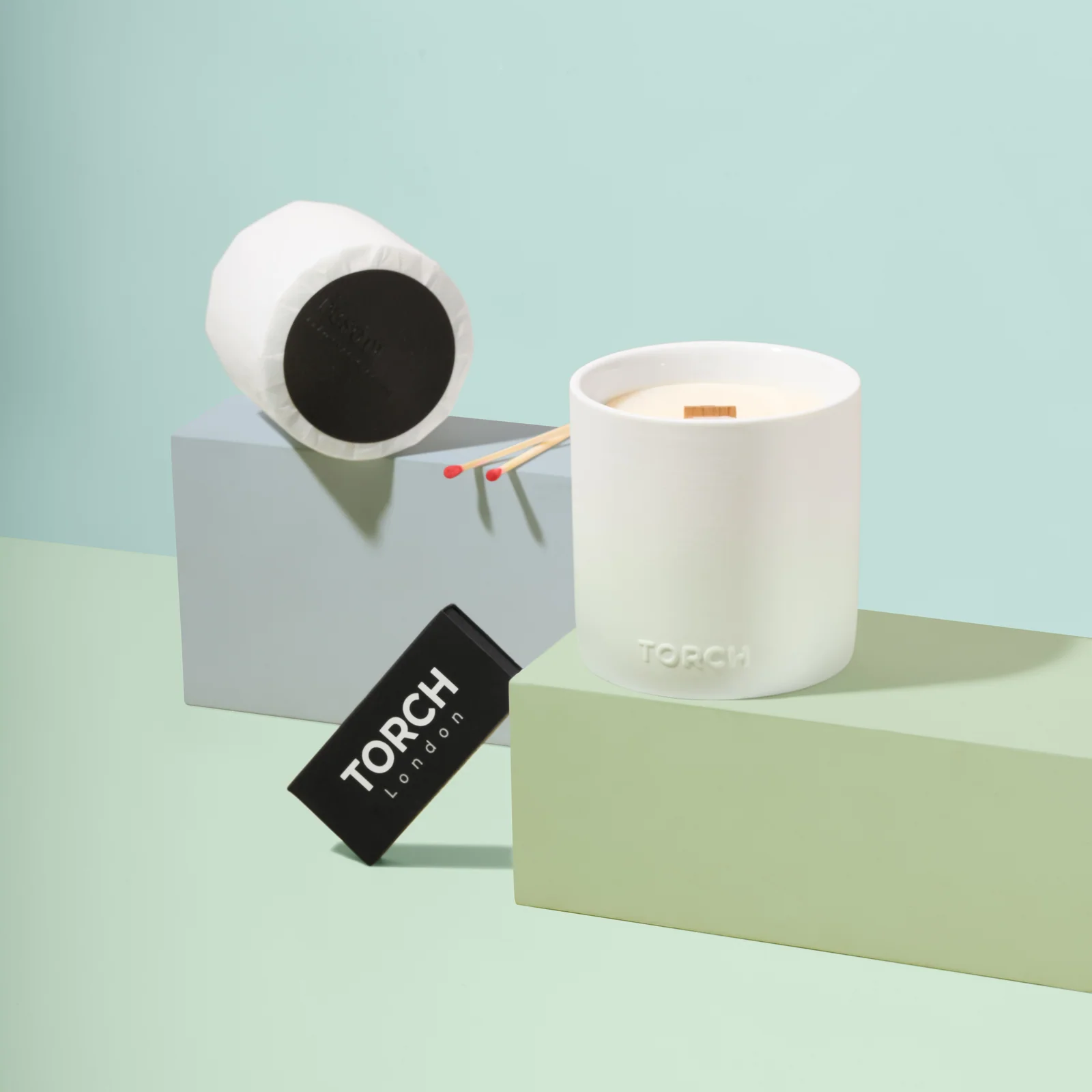 White Torch candle with a wrapped refill, matches and a black matchbox on pastel green and blue platforms.