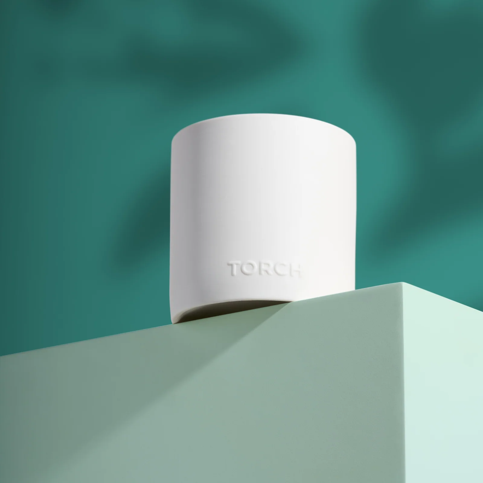 Close-up of a white Torch candle vessel on a pale green plinth against teal with soft shadows.