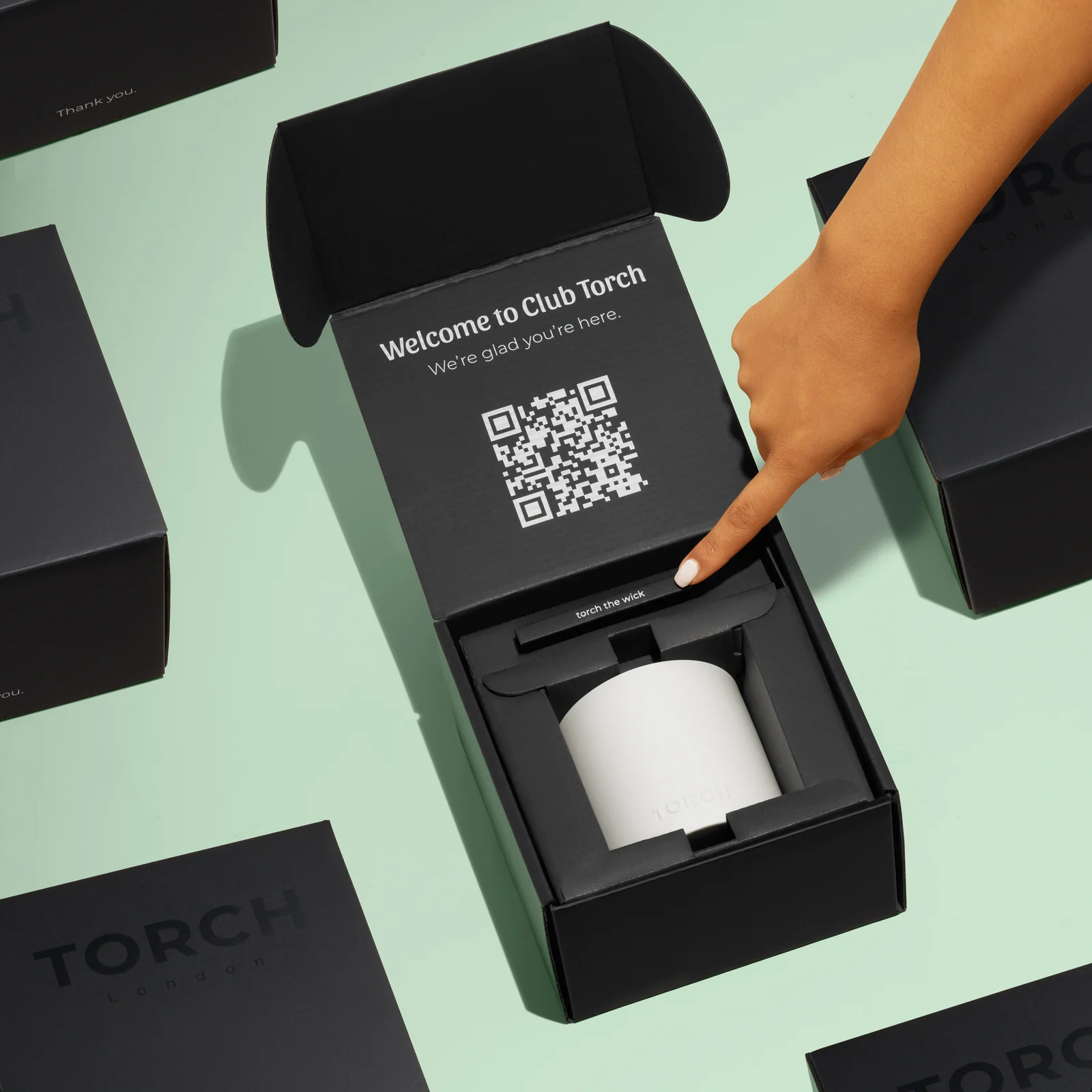 Hand pointing inside an open black Torch candle box with a QR code on the lid, surrounded by closed boxes on green.