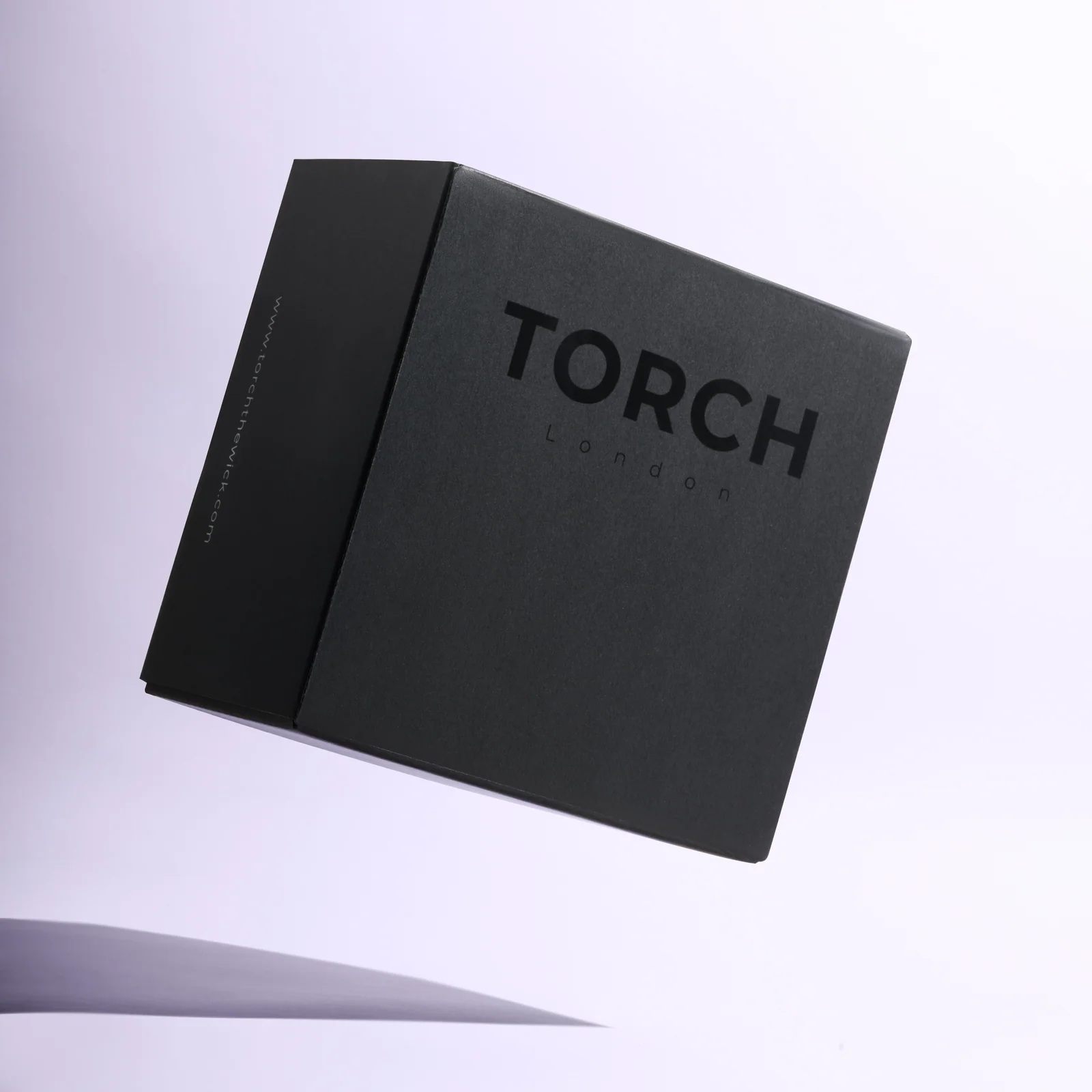 Black Torch candle box suspended at an angle above a soft shadow against lilac.