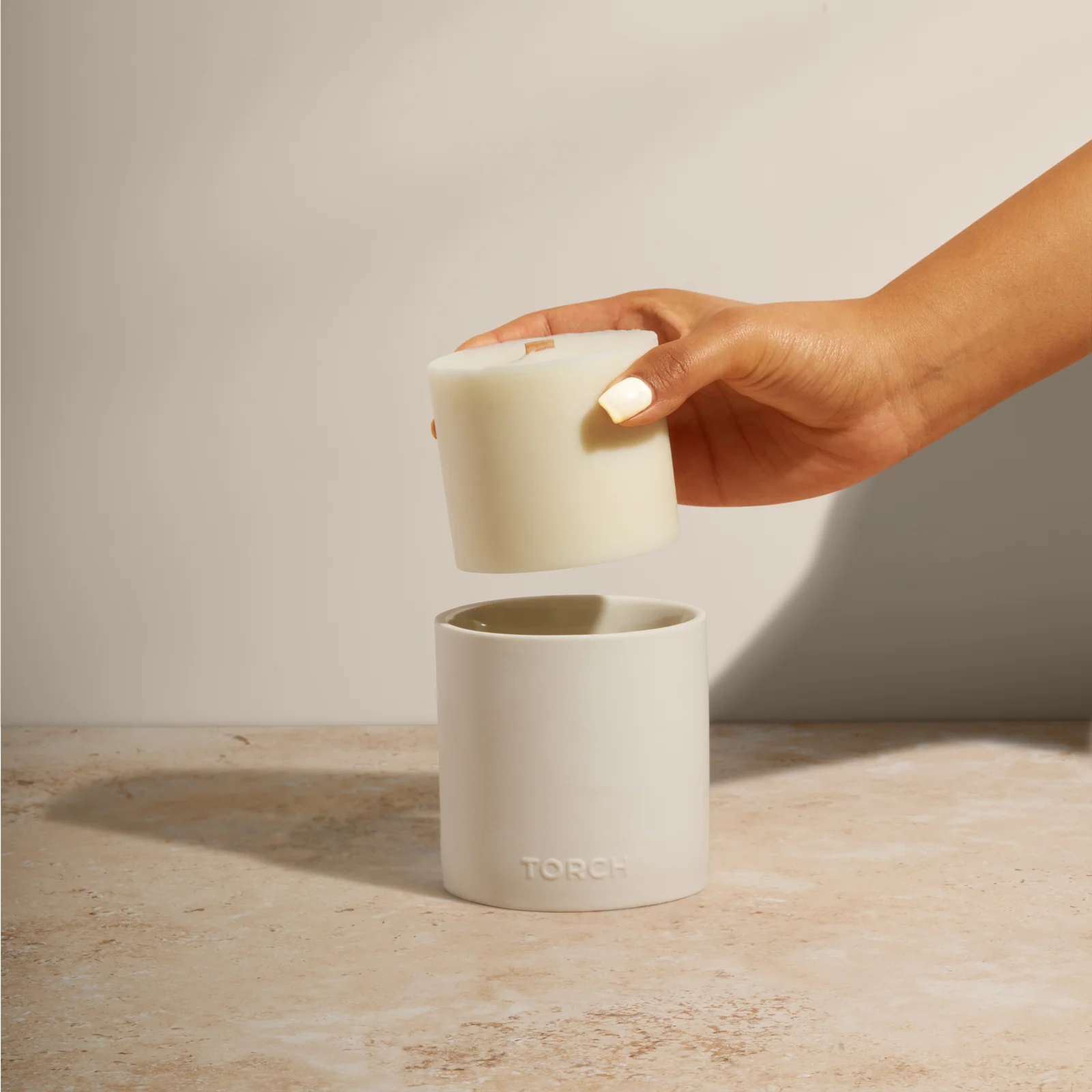 Hand holding a wax candle refill above an empty white Torch vessel on a stone surface against beige.