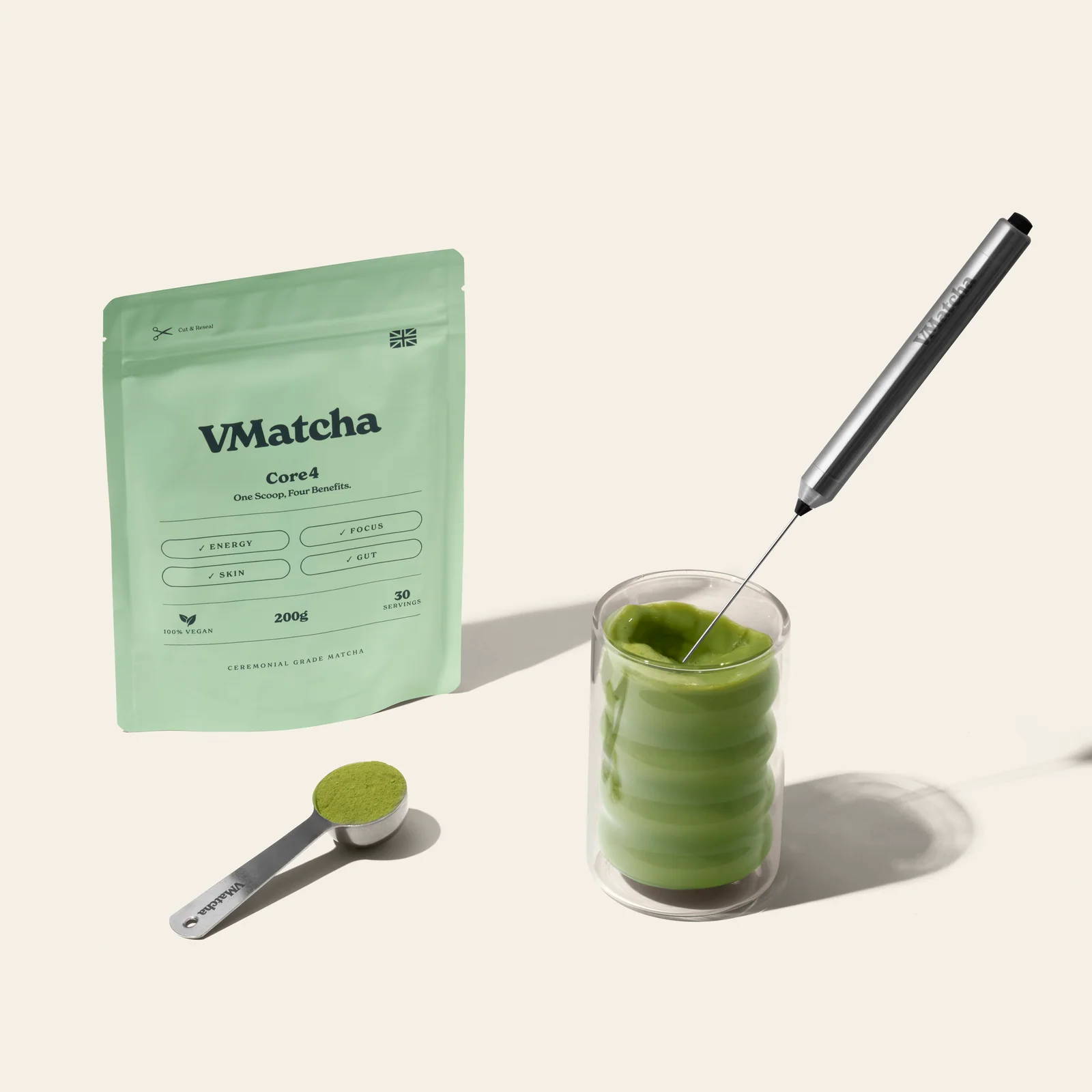 VMatcha Core4 pouch beside a glass of matcha, handheld whisk and scoop on a cream surface.