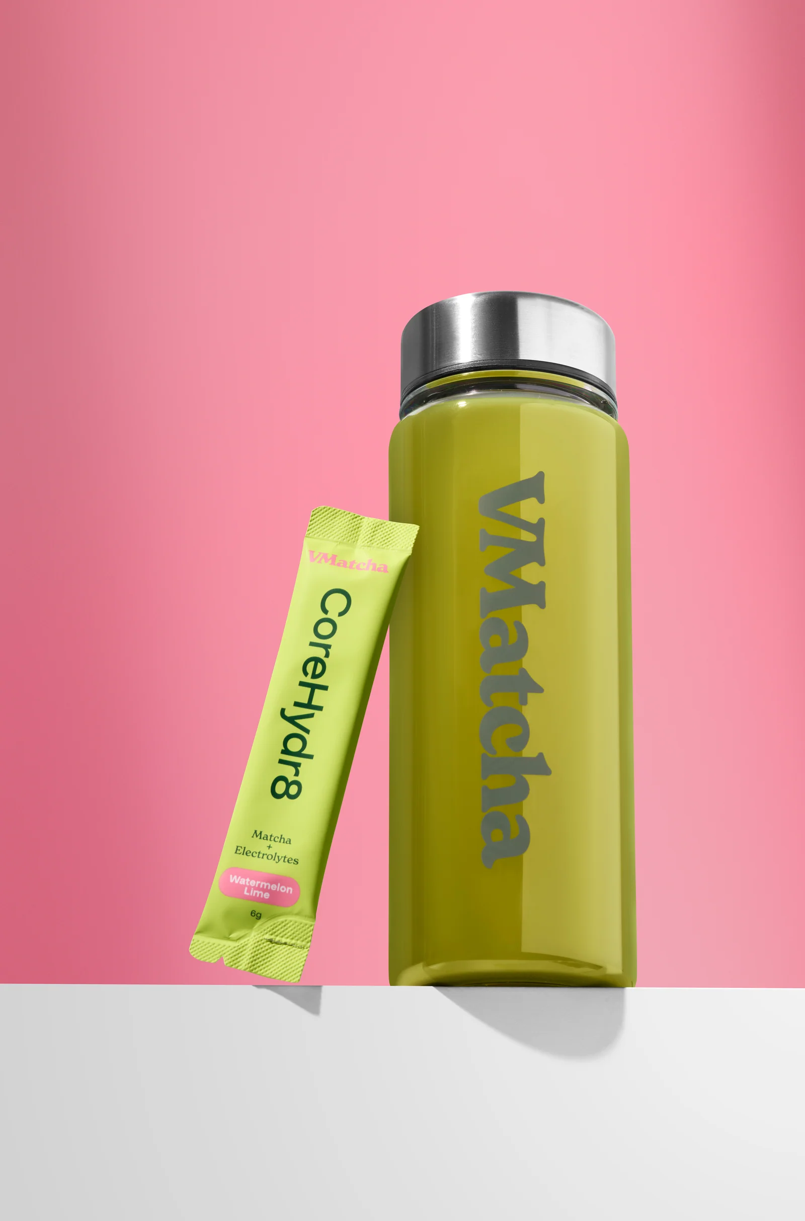 VMatcha CoreHydr8 sachet beside a bottle of green drink on a white ledge against pink.