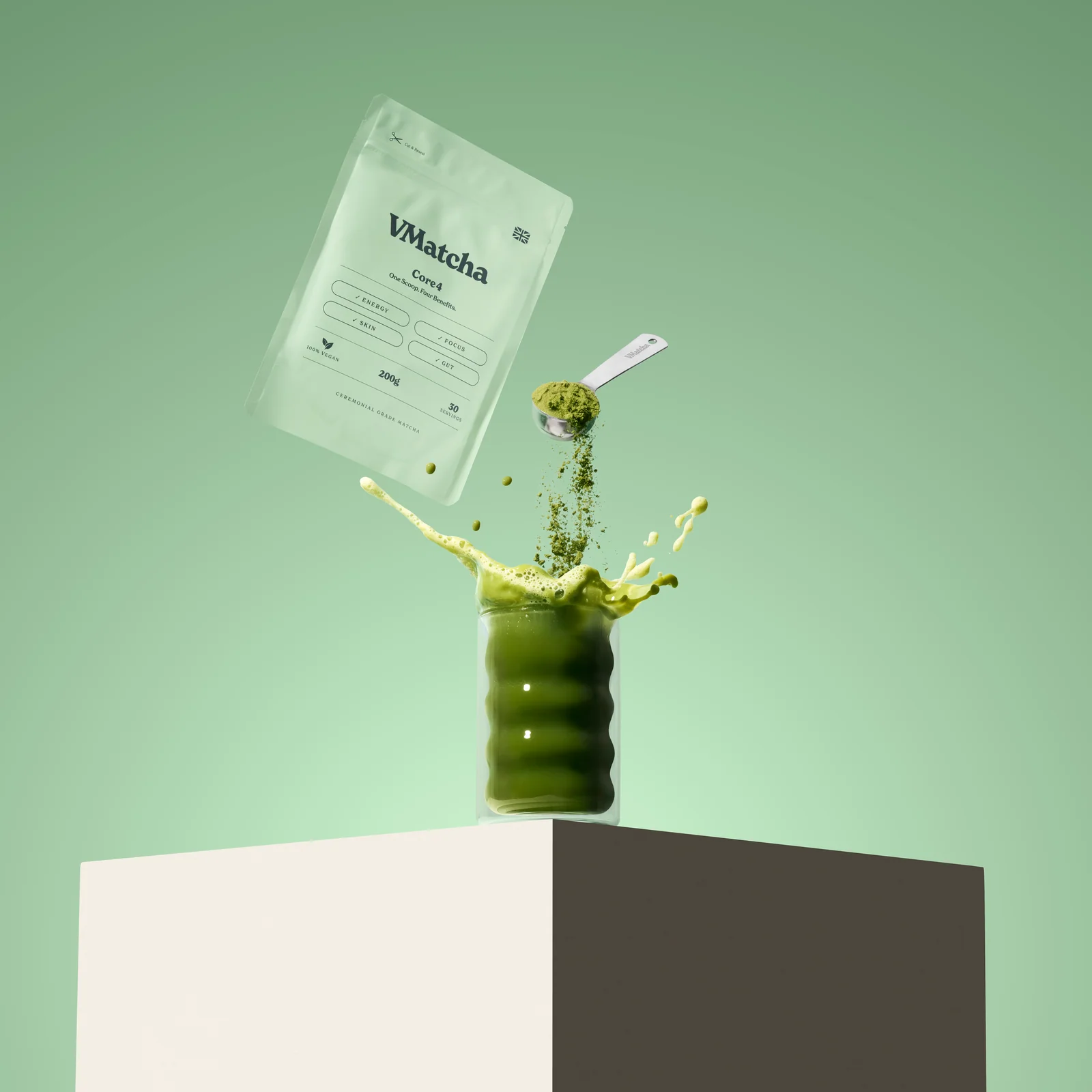 VMatcha Core4 pouch beside a scoop pouring matcha powder into a splashing green drink on a pale green set.