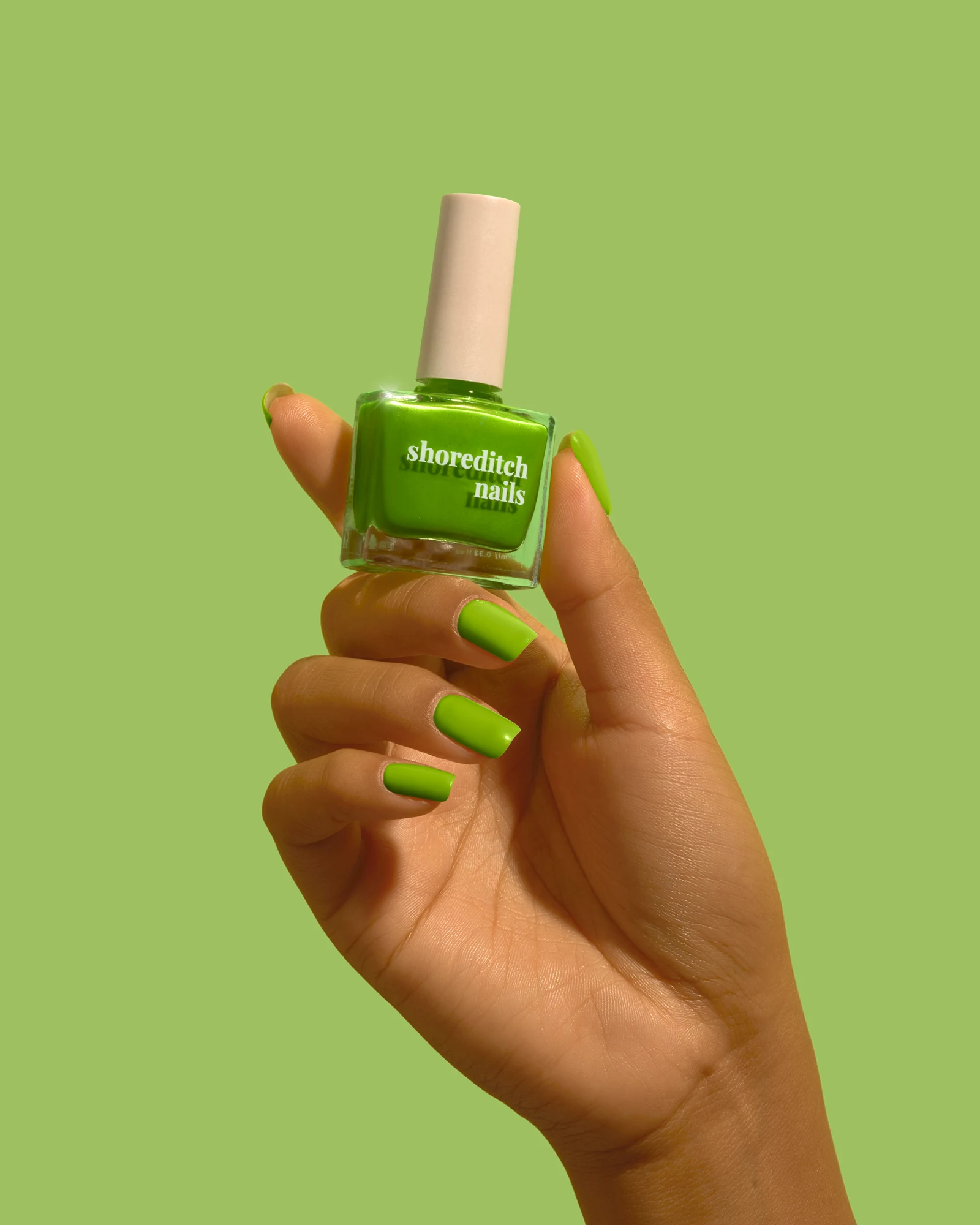 Hand with green nail polish holding a green Shoreditch Nails bottle against a green background.