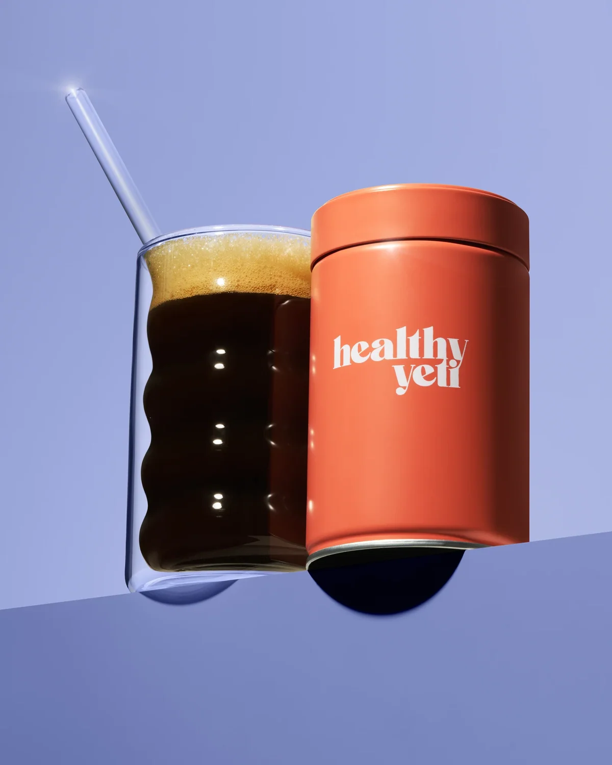 Orange Healthy Yeti jar beside a glass of dark coffee against lilac.