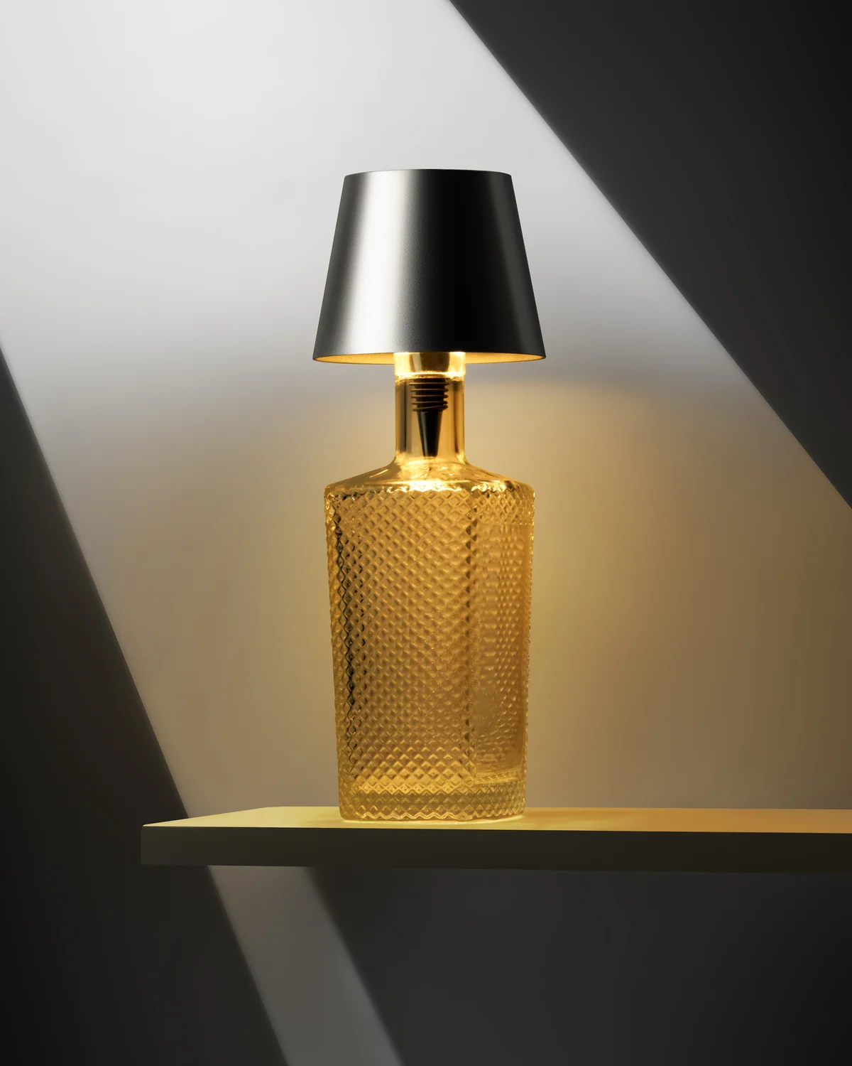 Textured amber-glass Liquor Lit bottle lamp with a black shade, illuminated against grey.