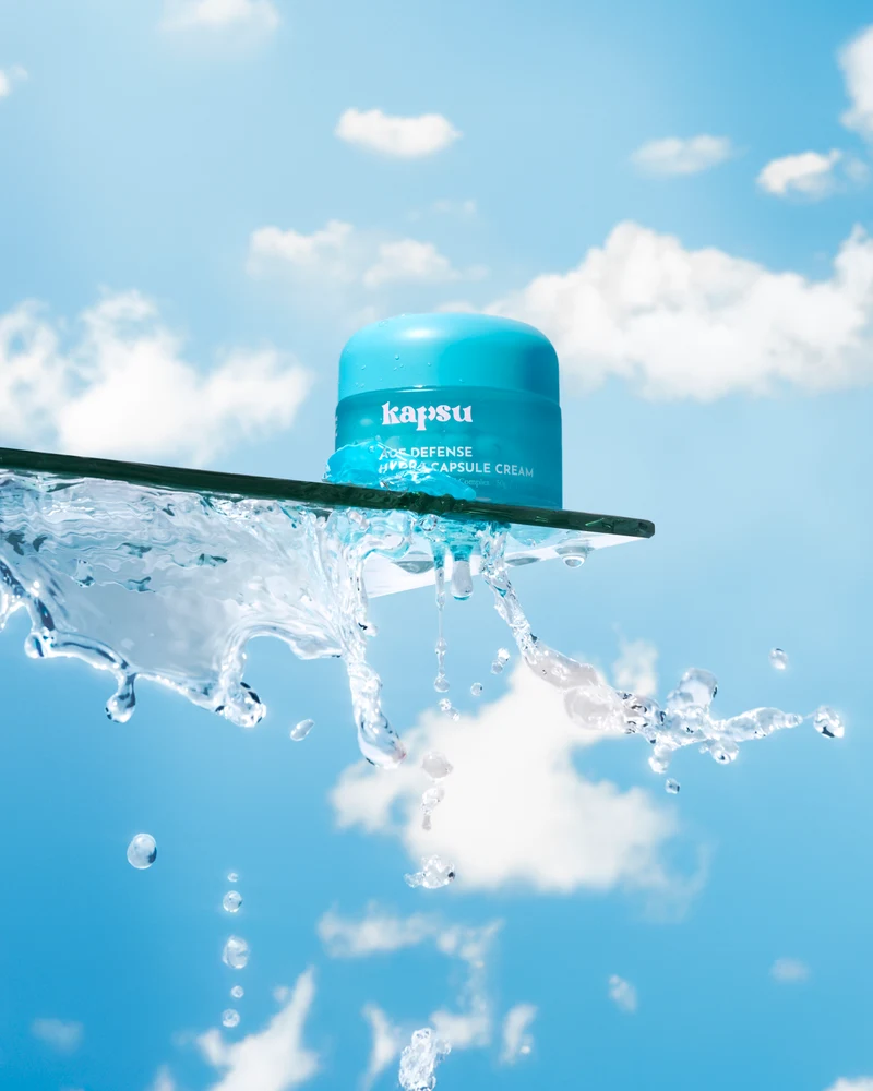 Turquoise Kapsu skincare jar on a glass shelf with splashing water against a blue sky.